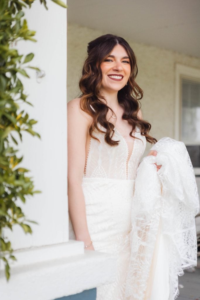 A woman in a white wedding dress stands near a wall at The Barns at Hamilton Station, smiling and holding part of her dress. She has long, curled hair and is positioned next to a leafy plant, as she enjoys the final moments of preparation for her wedding.