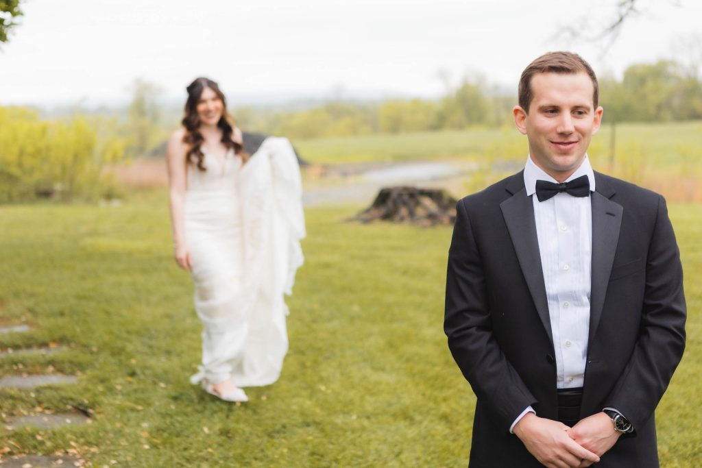 A bride in a white dress walks towards a groom in a black suit and bow tie, standing outdoors on the grassy area of The Barns at Hamilton Station, where their wedding preparations have reached a beautiful climax.