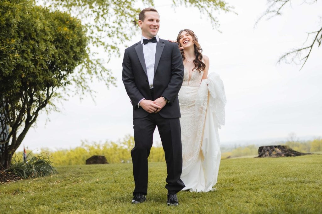 A bride in a white dress and groom in a black tuxedo stand on lush grass, smiling and laughing, with trees and an overcast sky in the background at The Barns at Hamilton Station.