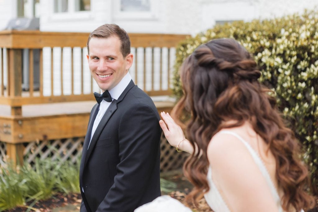 A man in a black suit and bow tie smiles as a woman with long, wavy hair in a white dress touches his shoulder. They are outdoors near the wooden deck at The Barns at Hamilton Station, the perfect spot for wedding preparation.