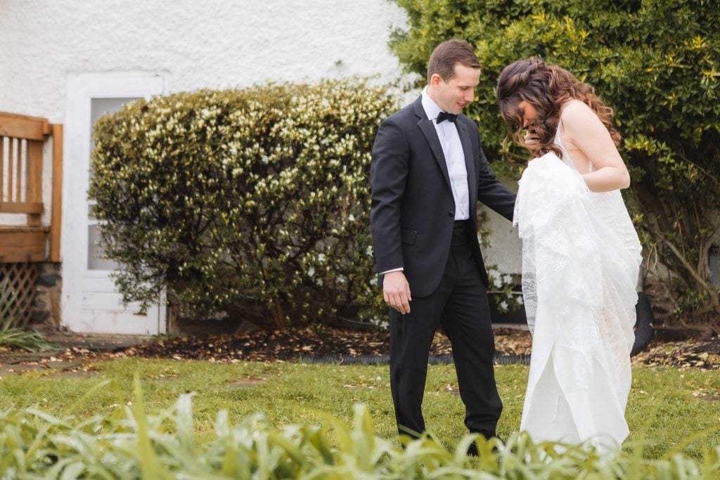 A man in a black suit and a woman in a white dress stand on the grass at The Barns at Hamilton Station, looking down. Shrubs and a white wall are in the background, hinting at careful wedding preparation.