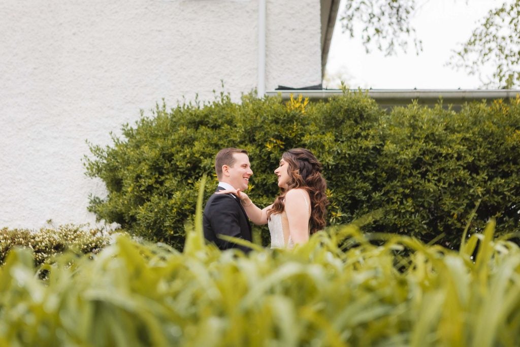 A couple, dressed formally in wedding attire, stand facing each other outside in front of a large green shrub with The Barns at Hamilton Station building in the background, capturing a moment of wedding preparation.