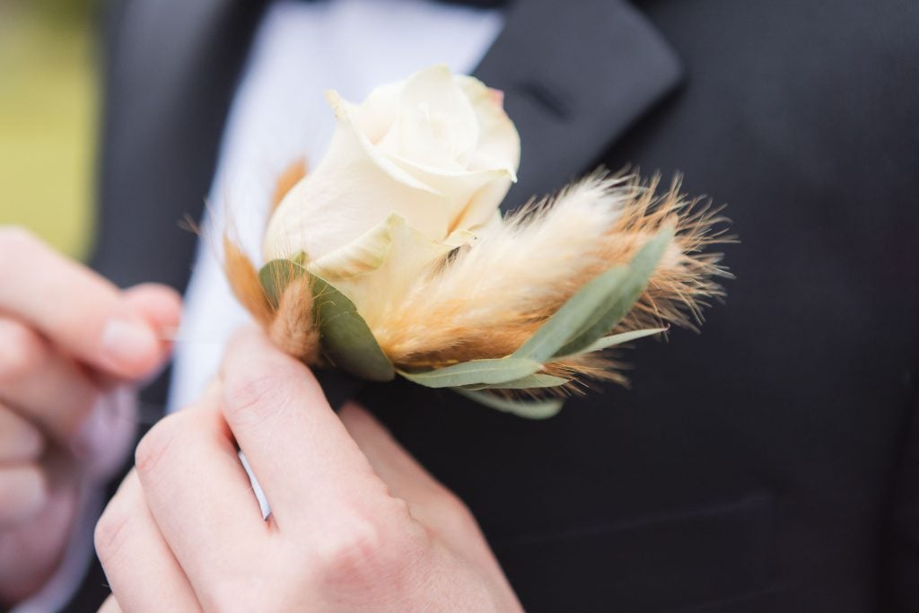 A person wearing a suit adjusts a white rose boutonniere, intricately decorated with dried grasses and greenery, paying attention to the details at The Barns at Hamilton Station wedding.