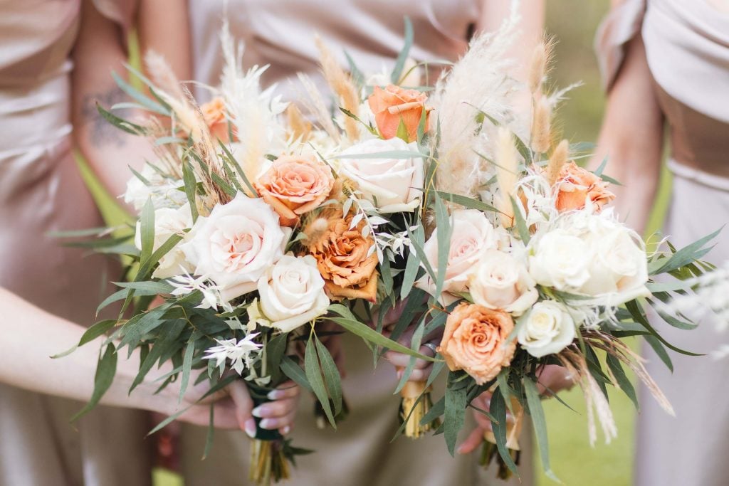 Three individuals in beige dresses hold elegant bouquets featuring white and peach roses, green foliage, and decorative grasses—a beautifully detailed scene at The Barns at Hamilton Station wedding.