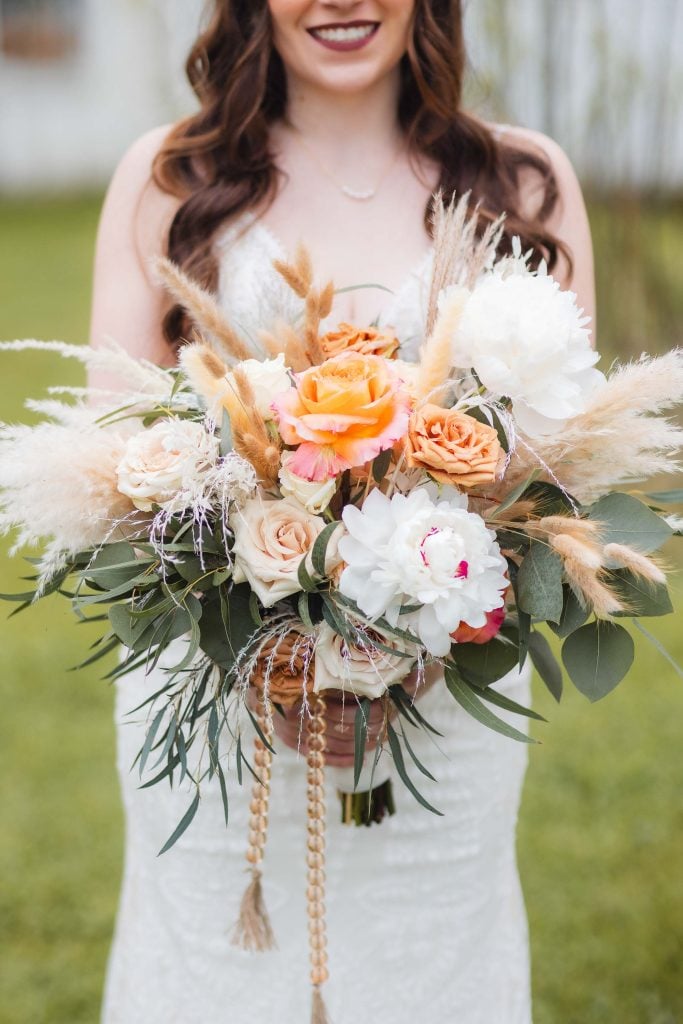 A woman in a white dress holds a bouquet with a mix of roses, peonies, greenery, and pampas grass at The Barns at Hamilton Station, perfect for wedding details.