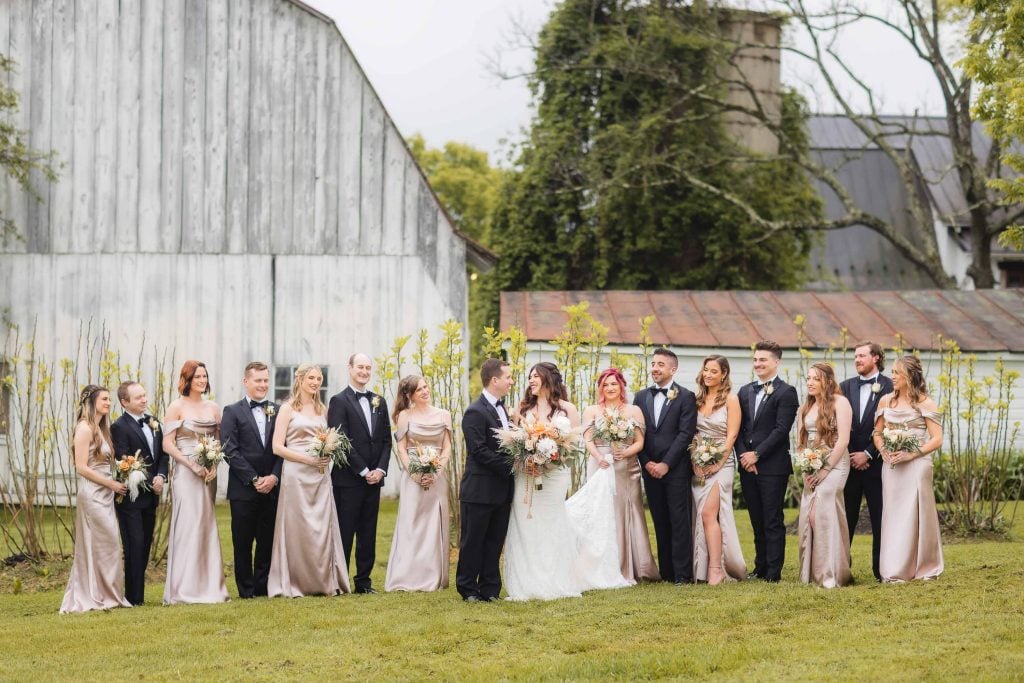 A wedding party stands outdoors in front of rustic buildings at The Barns at Hamilton Station. The group, resembling a picturesque portrait, includes bridesmaids in light dresses and groomsmen in dark suits, with the bride and groom in the center holding bouquets amidst a scenic vineyard backdrop.