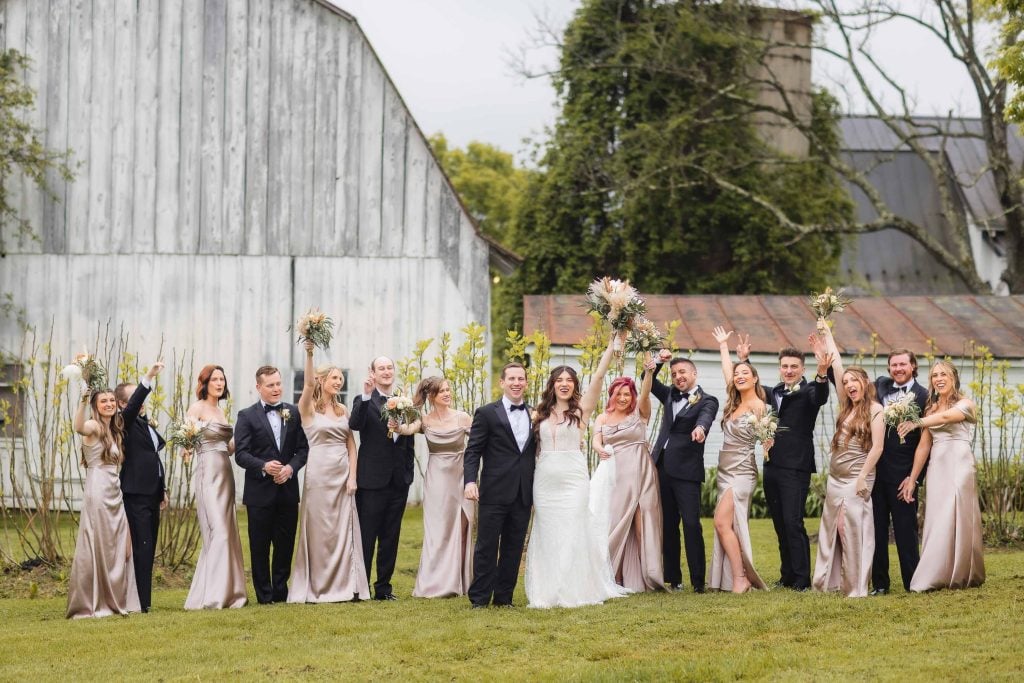 A wedding party stands in front of a rustic barn at a charming vineyard, with the bride and groom in the center. Bridesmaids in matching dresses and groomsmen in suits raise their arms and bouquets in celebration, creating a perfect portrait of joy and unity.