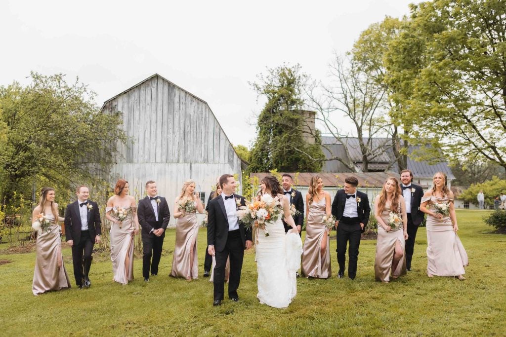 A wedding party stands outdoors in front of a rustic barn, with the bride and groom holding hands and surrounded by bridesmaids in light gowns and groomsmen in dark suits, creating a picturesque portrait akin to a scene from a charming vineyard celebration.