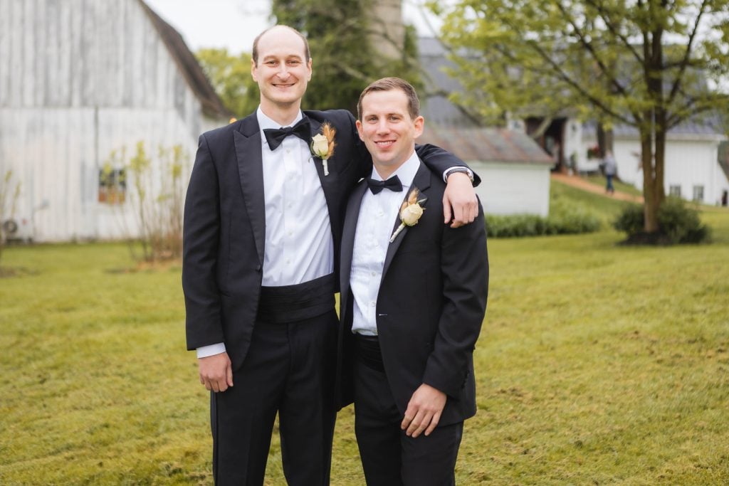 Two men in black tuxedos stand outdoors on a grassy lawn, one with his arm around the other's shoulder. The Barns at Hamilton Station serve as their elegant backdrop, with a white barn and trees framing the scene, capturing the essence of a vineyard wedding.