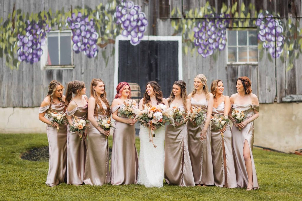 A bride stands with her bridesmaids, all wearing matching light gold dresses, in front of an old wooden barn with painted grapevines. They are holding floral bouquets and smiling, capturing a perfect wedding portrait amidst this charming vineyard setting.