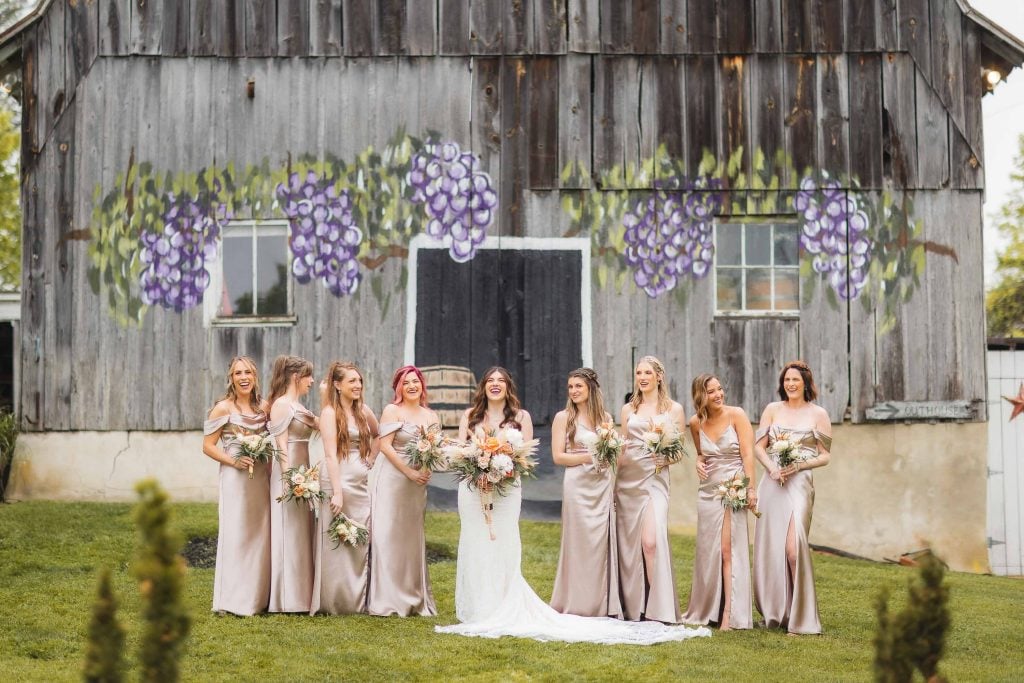 A wedding portrait captures a bride and her eight bridesmaids in matching beige dresses standing in front of a wooden barn adorned with painted grapes, evoking a vineyard charm. Each bridesmaid holds a bouquet, adding to the picturesque scene.