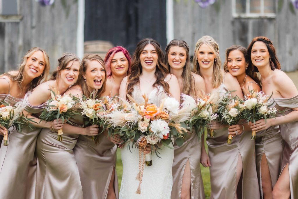 A bride in a white dress stands smiling with eight bridesmaids in taupe dresses, holding bouquets of flowers in front of the rustic backdrop of The Barns at Hamilton Station.