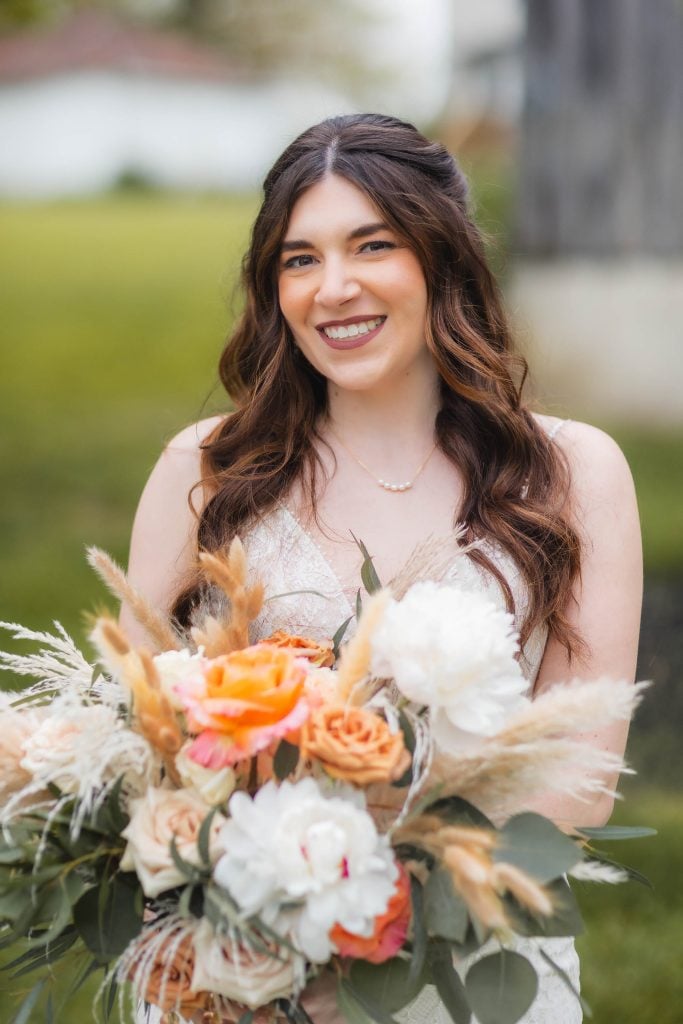 A woman with long brown hair smiles while holding a bouquet of orange, white, and beige flowers. She is wearing a white dress and a simple necklace. The background is an outdoor wedding amidst lush greenery and vineyard views.