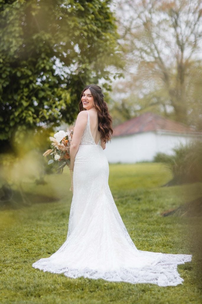 A bride in a white wedding dress stands outdoors on grass, holding a bouquet and looking over her shoulder with a smile. Trees, a building, and a vineyard are in the background.