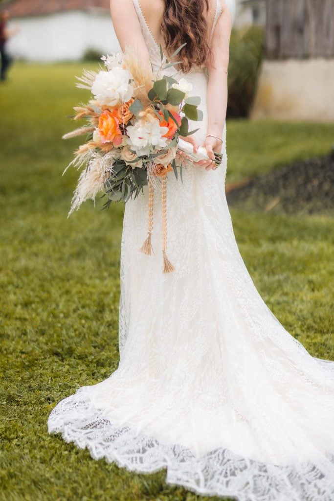 A woman in a white lace wedding dress holds a bouquet of flowers while standing on grass at The Barns at Hamilton Station. Her back is turned to the camera, capturing a serene portrait against the backdrop of the vineyard.