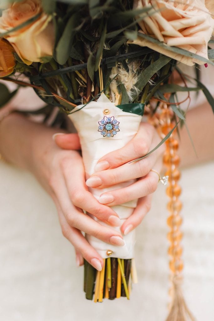 Closeup of hands holding a bouquet of flowers wrapped in white fabric, adorned with a jeweled brooch. The hands have neatly manicured nails, and one wears a diamond ring. The intricate details make it perfect for a wedding at The Barns at Hamilton Station.