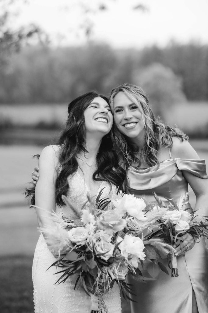 Two women are standing outdoors at The Barns at Hamilton Station, smiling and holding bouquets. One is wearing a white dress, and the other is in an offshoulder dress. The background is blurred, showing trees and a field, adding to the charm of this vineyard wedding scene.