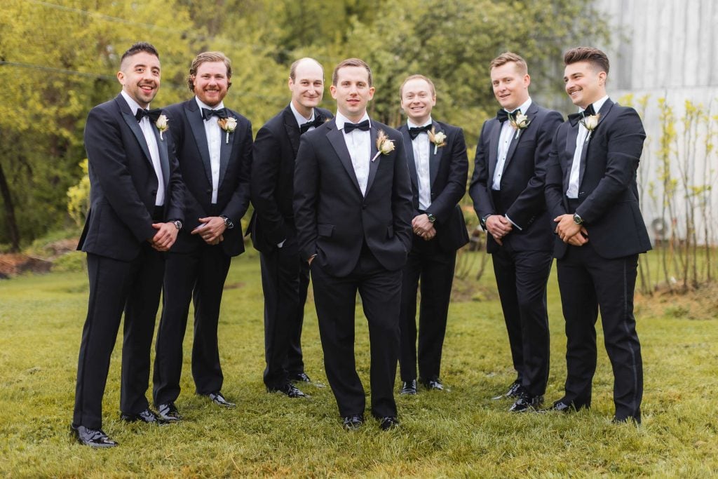 A group of seven men dressed in black tuxedos with bow ties and boutonnières stand outdoors on the grass at The Barns at Hamilton Station, with trees and a building in the background, celebrating a beautiful wedding day.