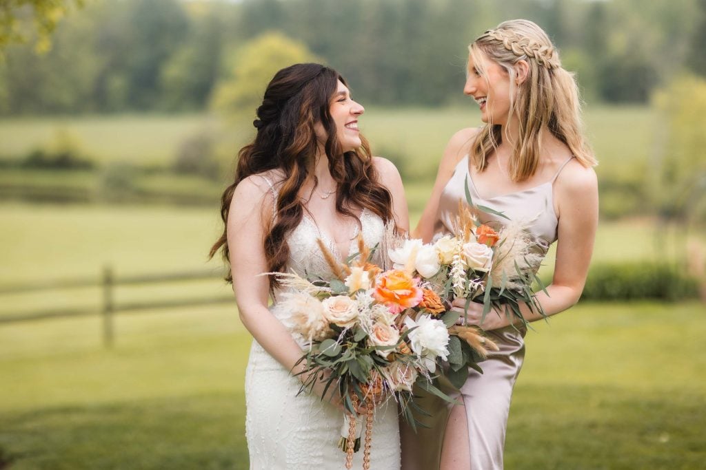 Two women smiling at each other outdoors, each holding a bouquet of flowers. One is wearing a white dress, and the other is in a lightcolored dress. A blurred green field in the background hints at a picturesque vineyard, creating an enchanting wedding portrait.