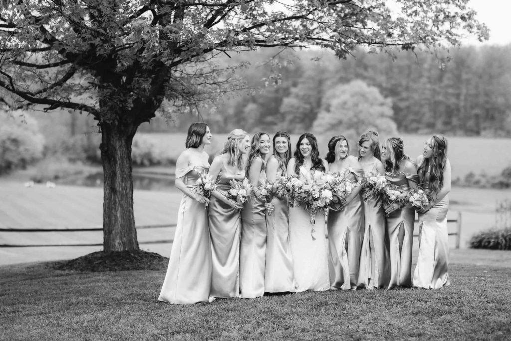 A group of bridesmaids in matching lightcolored dresses hold bouquets and smile together under a tree in an outdoor setting, creating the perfect wedding portrait at The Barns at Hamilton Station.