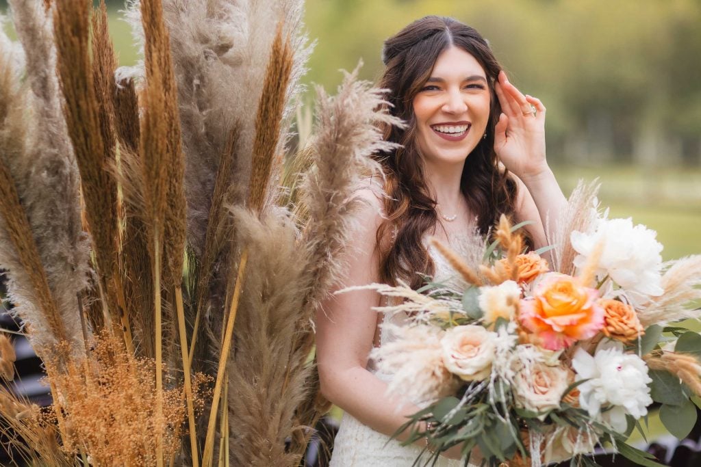 A smiling woman in a white dress holds a bouquet of flowers, standing beside tall, feathery plants in an outdoor vineyard setting.