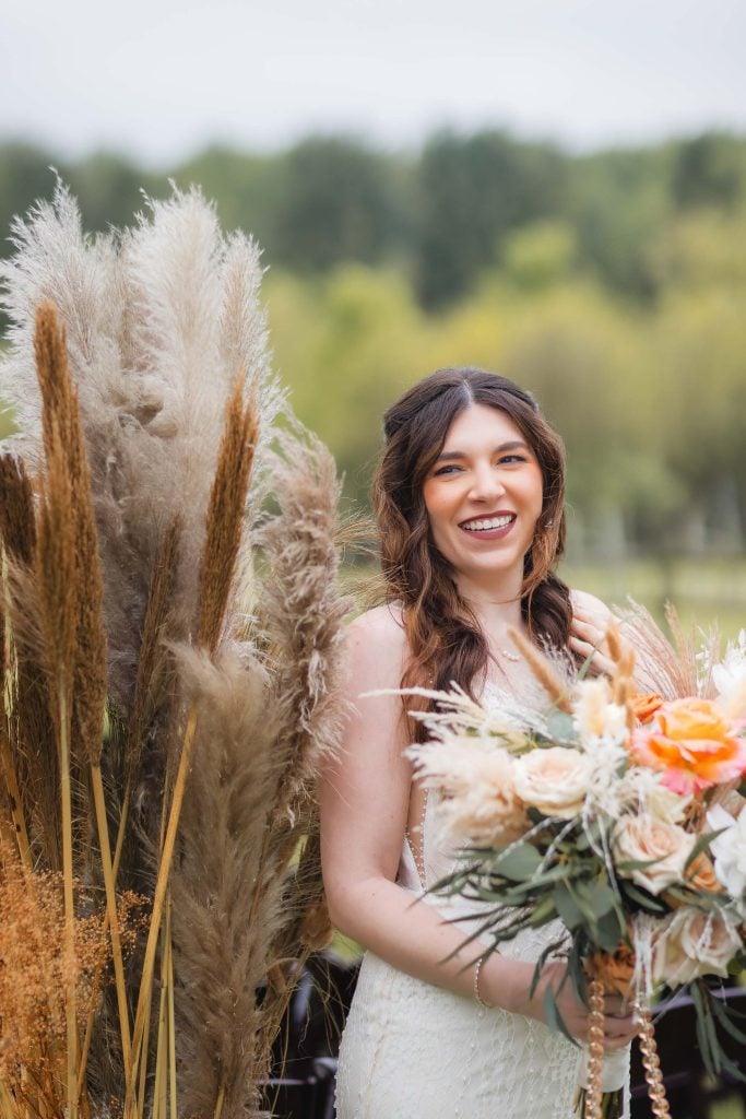 A smiling woman in a white dress holding a bouquet of flowers stands outdoors near tall ornamental grasses, capturing the essence of a picturesque wedding portrait amid vineyard scenery.