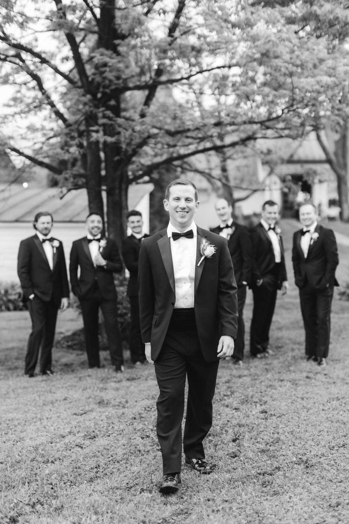 A groom in a tuxedo walks towards the camera with groomsmen standing behind him in a picturesque vineyard at The Barns at Hamilton Station.