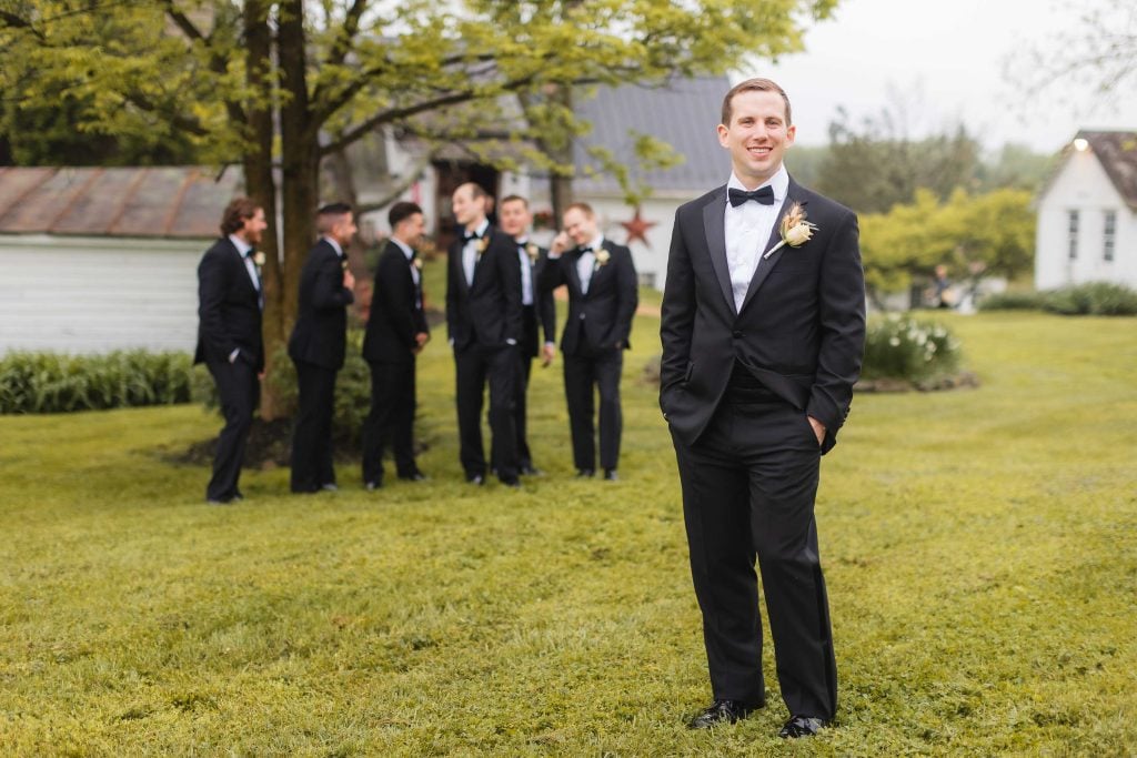 A groom in a black tuxedo stands on a grassy lawn, smiling at the camera. In the background, groomsmen in black tuxedos gather near a tree and a rustic building, creating a picturesque wedding portrait that could easily be set in an elegant vineyard.