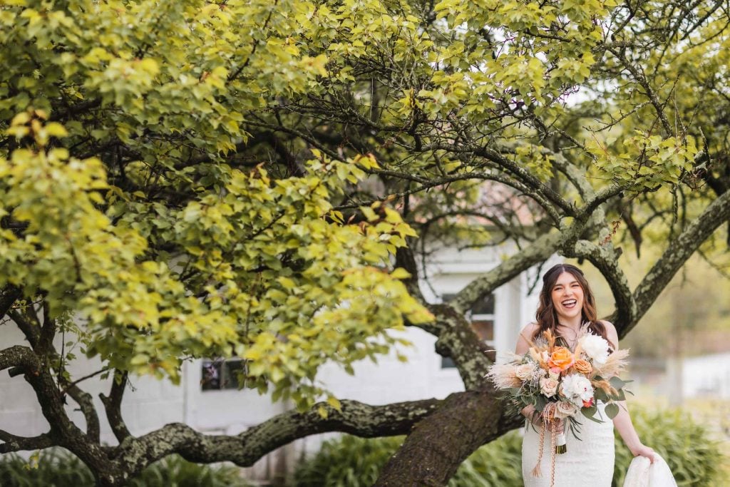 A bride in a white dress stands outdoors at The Barns at Hamilton Station, smiling and holding a bouquet of flowers. Behind her, a large, leafy tree completes the picturesque wedding portrait.