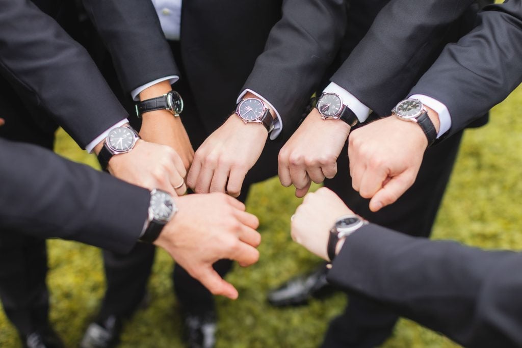 Eight people wearing black suits display their wristwatches, forming a circle with their hands extended inward. The setting is a wedding at The Barns at Hamilton Station, where every detail has been meticulously planned.