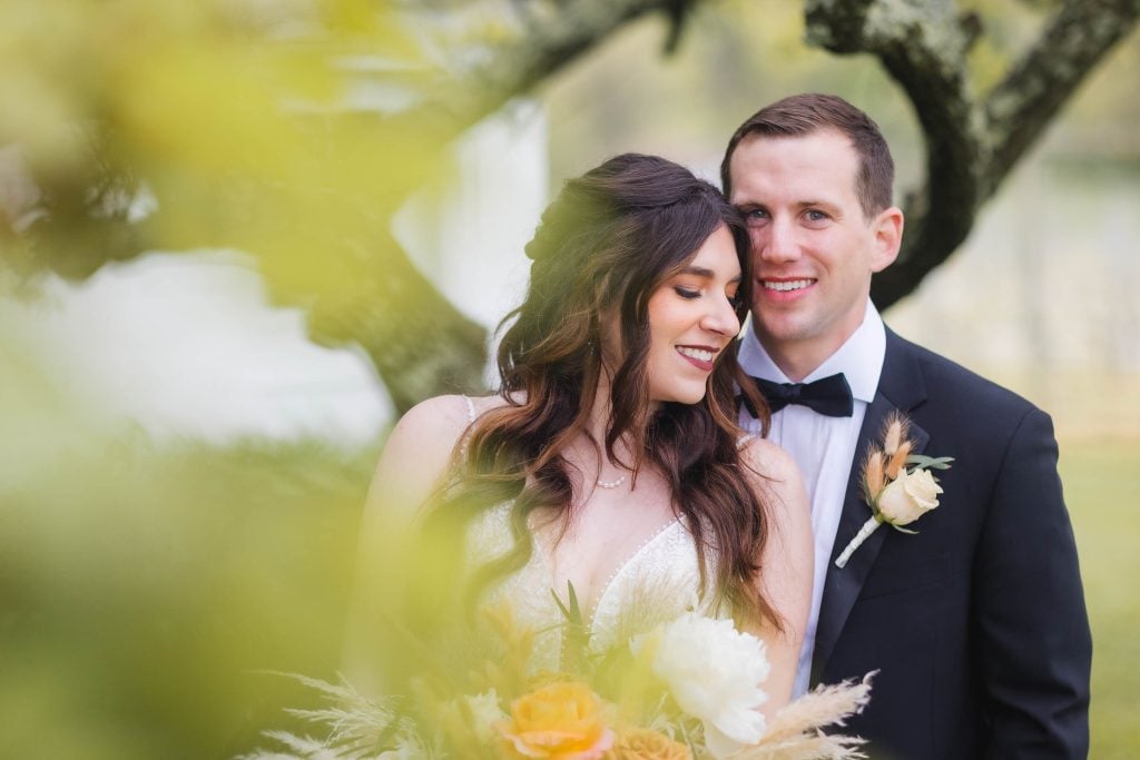 A couple, dressed in formal attire, stands close together outdoors in a vineyard. The woman wears a white dress and holds a bouquet, while the man is in a black suit with a bow tie and boutonniere. This wedding portrait captures their elegant simplicity amidst the lush vines.