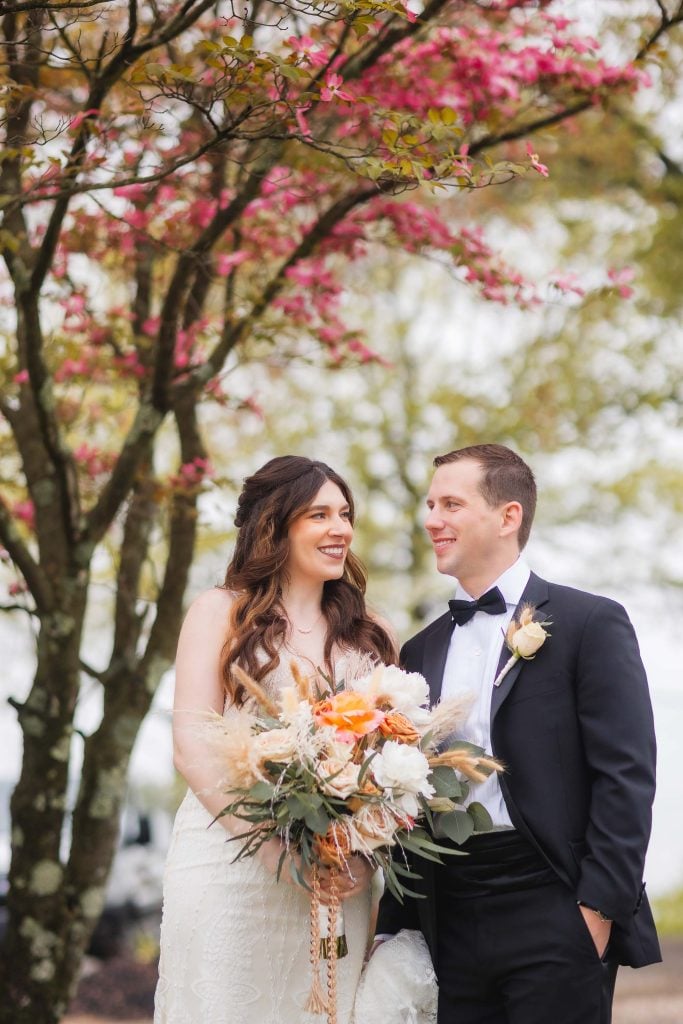 A bride and groom smile at each other in front of a flowering tree at The Barns at Hamilton Station. The bride holds a bouquet of flowers, and the groom is dressed in a black suit with a bow tie, capturing the perfect wedding portrait.