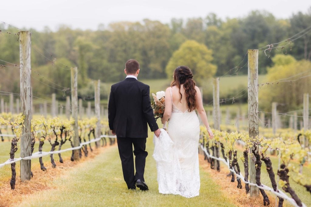 A bride and groom walk hand in hand down a vineyard aisle, wearing a white wedding dress and black suit respectively, surrounded by grapevines and greenery, creating a picturesque wedding portrait.