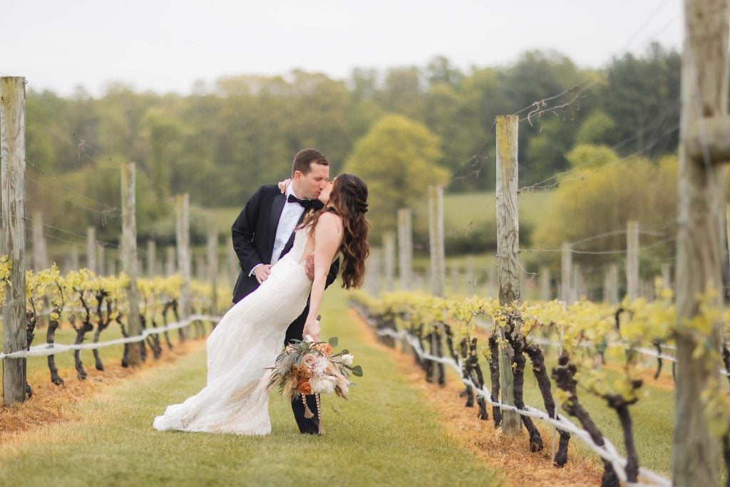 A portrait of a bride in a white dress and groom in a black suit sharing a kiss at The Barns at Hamilton Station, holding a bouquet amidst green vines, with trees in the background.