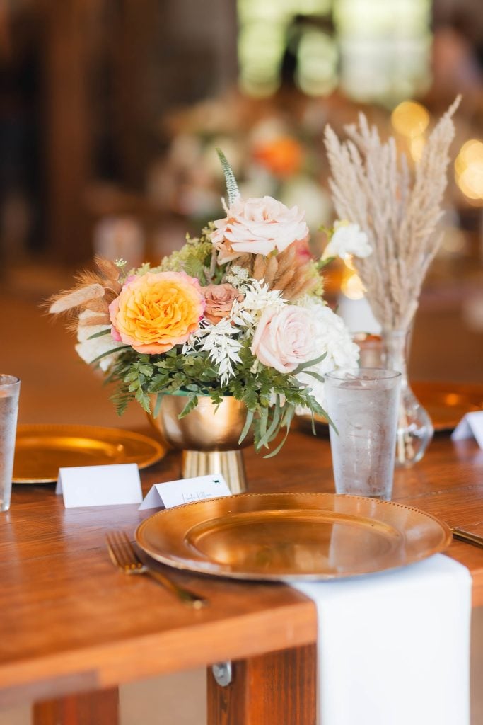 At The Barns at Hamilton Station, a wooden table set for a meal showcases exquisite details: a centerpiece of various flowers in a gold vase, gold chargers, empty glasses, and elegant place cards—perfect for an enchanting wedding.