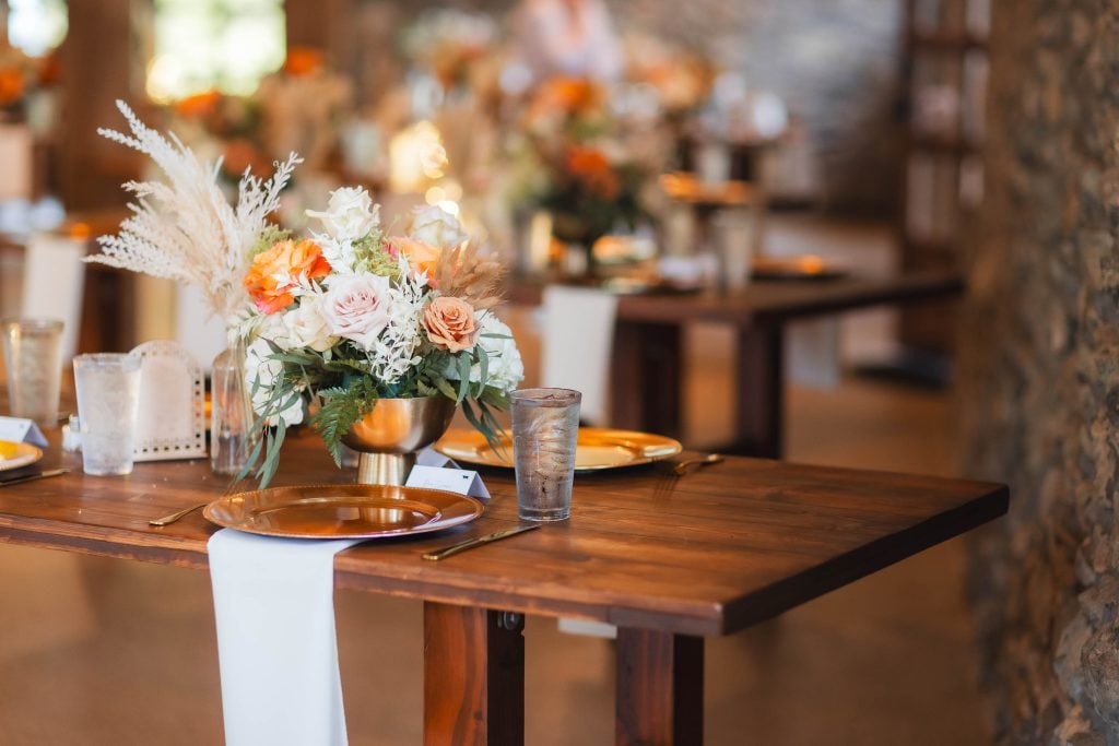 A wooden table is set with goldrimmed plates, clear glasses, and a flower arrangement featuring white, peach, and orange flowers. The wedding at The Barns at Hamilton Station showcases such elegant details, with the background revealing more similarly decorated tables.