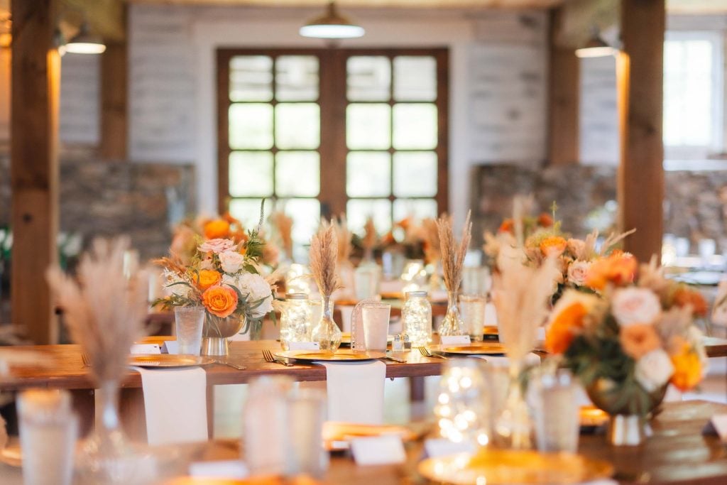 A rustic indoor venue, The Barns at Hamilton Station, features wooden tables adorned with floral centerpieces, candles, and wheat stalks. The details are enhanced by a large window in the background that floods the space with natural light.