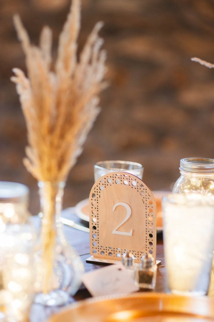 Closeup of a table setting at The Barns at Hamilton Station featuring a decorative table number 2, dried wheat stalks in a vase, and glass jars with string lights, creating a warm and rustic wedding ambiance with charming details.