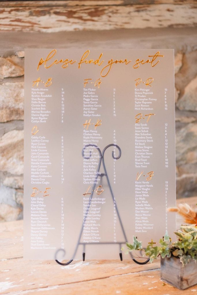 A seating chart with gold text titled "Please find your seat" displays guest names sorted by table number. The chart is placed on an easel against a stone wall backdrop with a small plant nearby, adding charming details perfect for a wedding at The Barns at Hamilton Station.