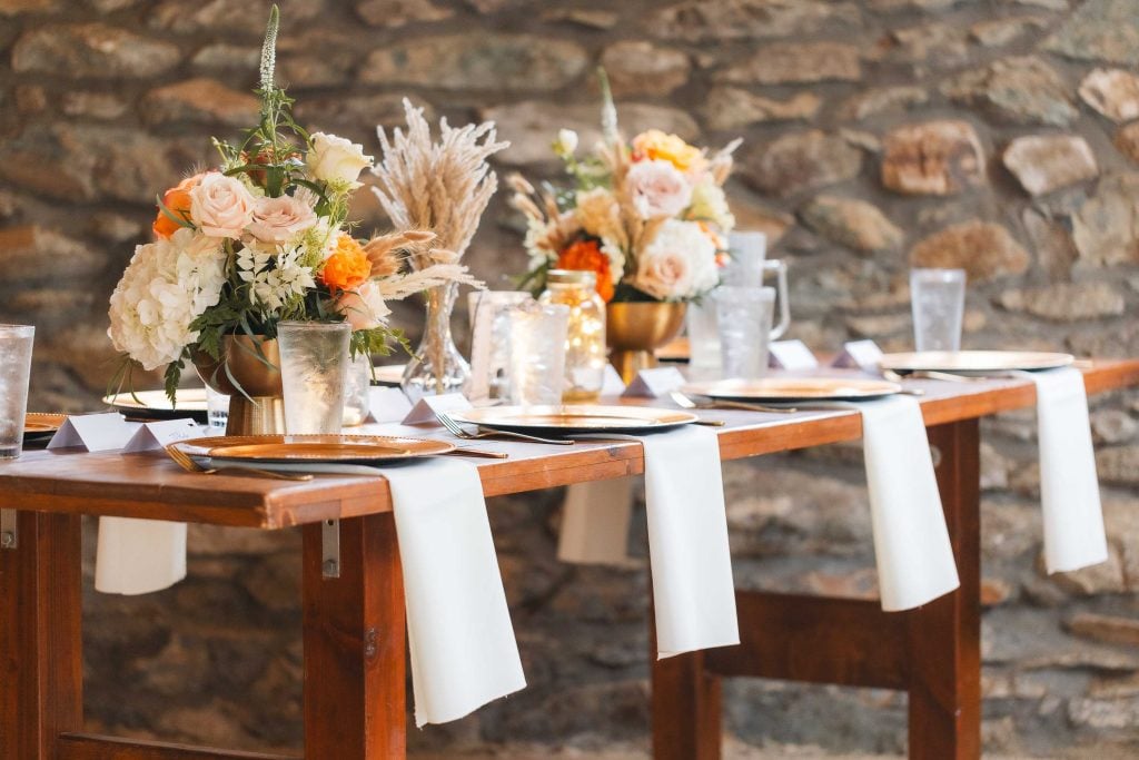 A wooden table set for a formal wedding at The Barns at Hamilton Station features white napkins, gold plates, glasses, and exquisite floral centerpieces, showcasing the meticulous details that make the occasion unforgettable.