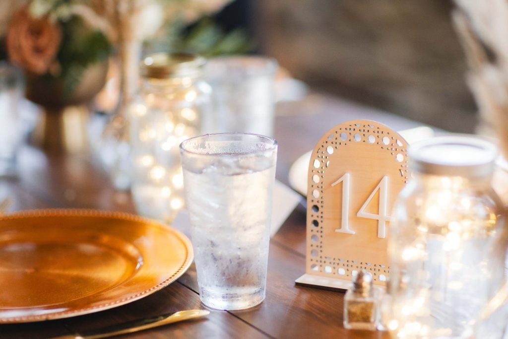 A decorated table at The Barns at Hamilton Station features a golden plate, a glass of water, a wooden table number 14 sign, and mason jars with lights. The wedding details create an enchanting ambiance.