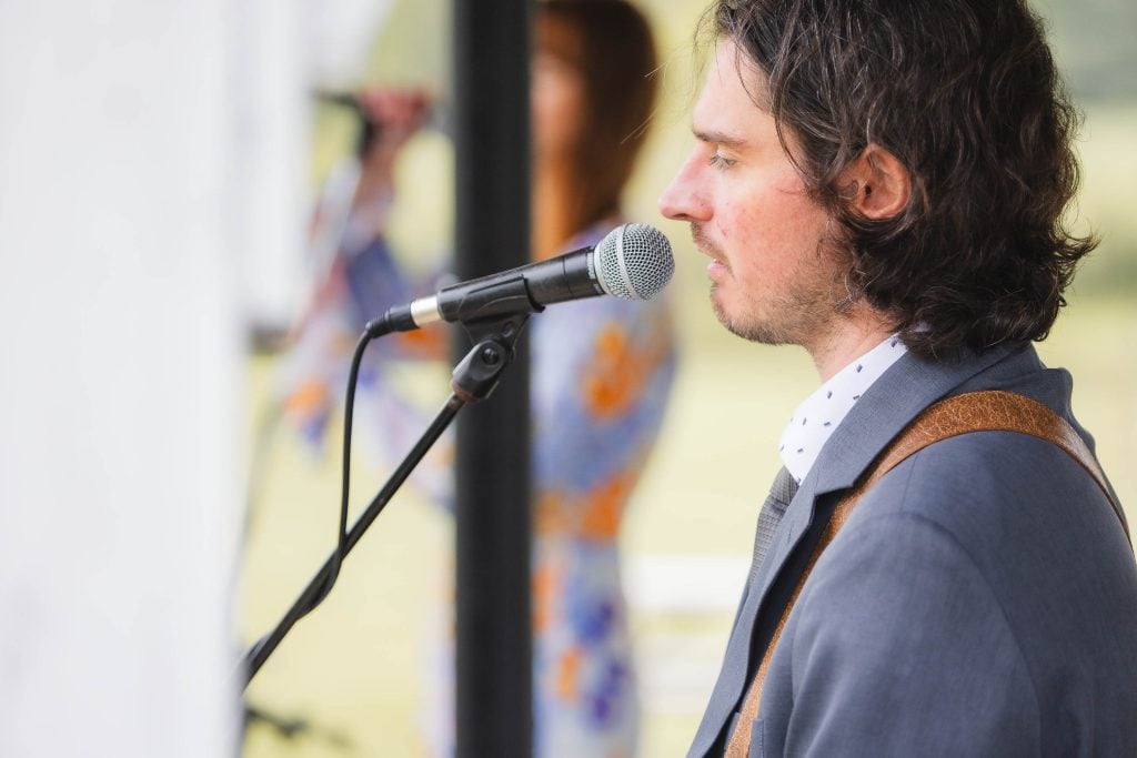 A man in a suit with long hair and beard sings into a microphone on stage at The Barns at Hamilton Station. A blurred figure appears in the background, adding a sense of mystery to the ceremony.