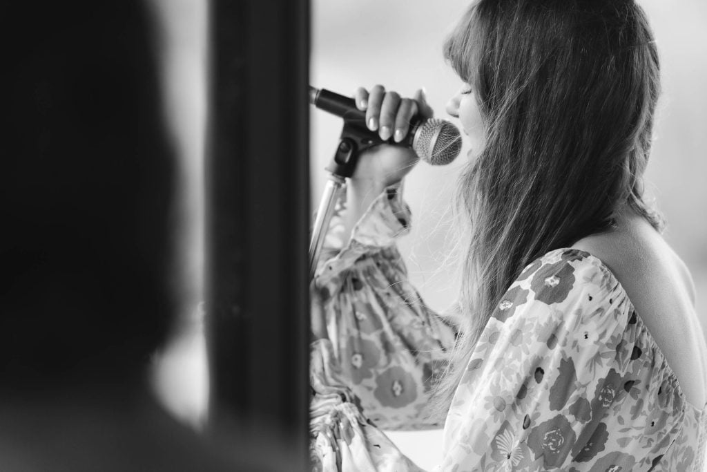 Black and white image of a person with long hair holding a microphone and singing, shown from a side angle at The Barns at Hamilton Station, with another blurry figure in the foreground.