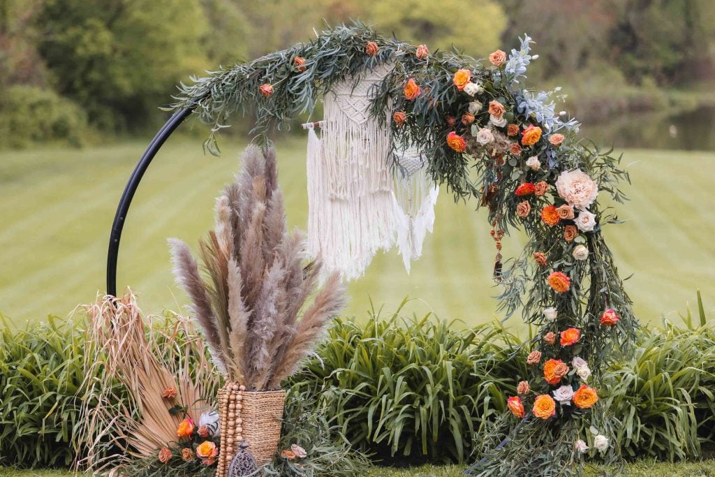 A semicircular floral arch with greenery, orange and white flowers, and a hanging macrame decor graces the backdrop at The Barns at Hamilton Station. Detailed with a basket of pampas grass and decorated leaves beside it, every element captures the essence of a perfect wedding setting outdoors.