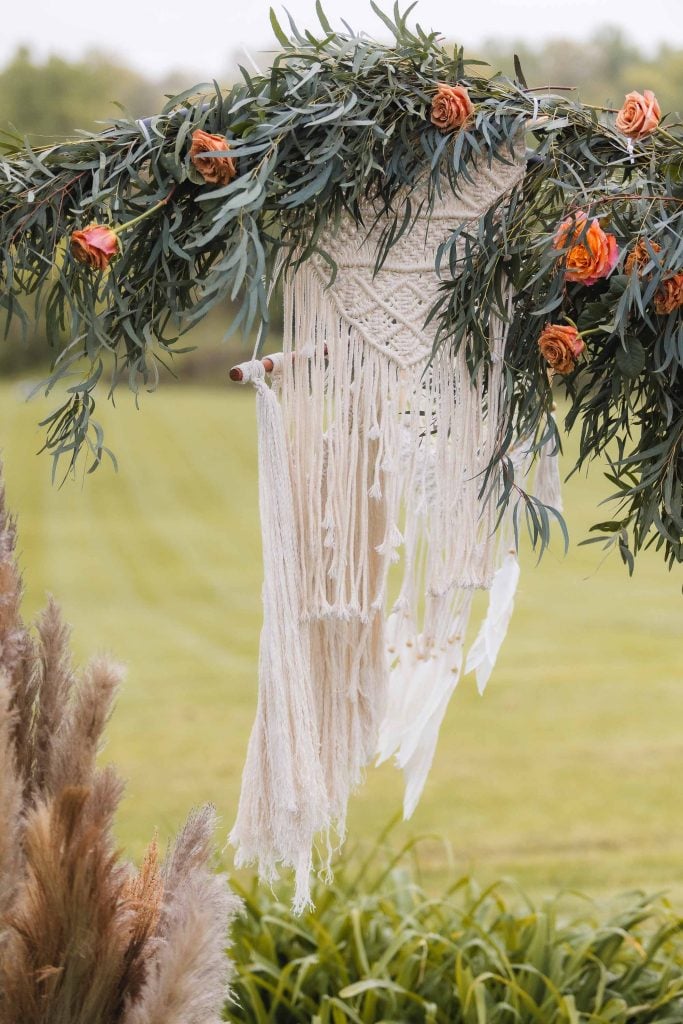 Macramé wall hanging adorned with foliage and orange flowers, suspended from a green natural backdrop, perfect for adding intricate details to your wedding decor at The Barns at Hamilton Station.