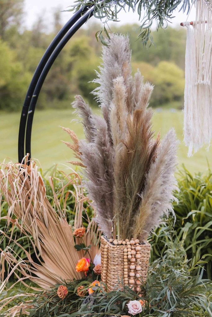 A decorative basket filled with various dried pampas grass arrangements, accented by a few pink roses, set outdoors at The Barns at Hamilton Station with lush green foliage in the background, perfect for highlighting wedding details.