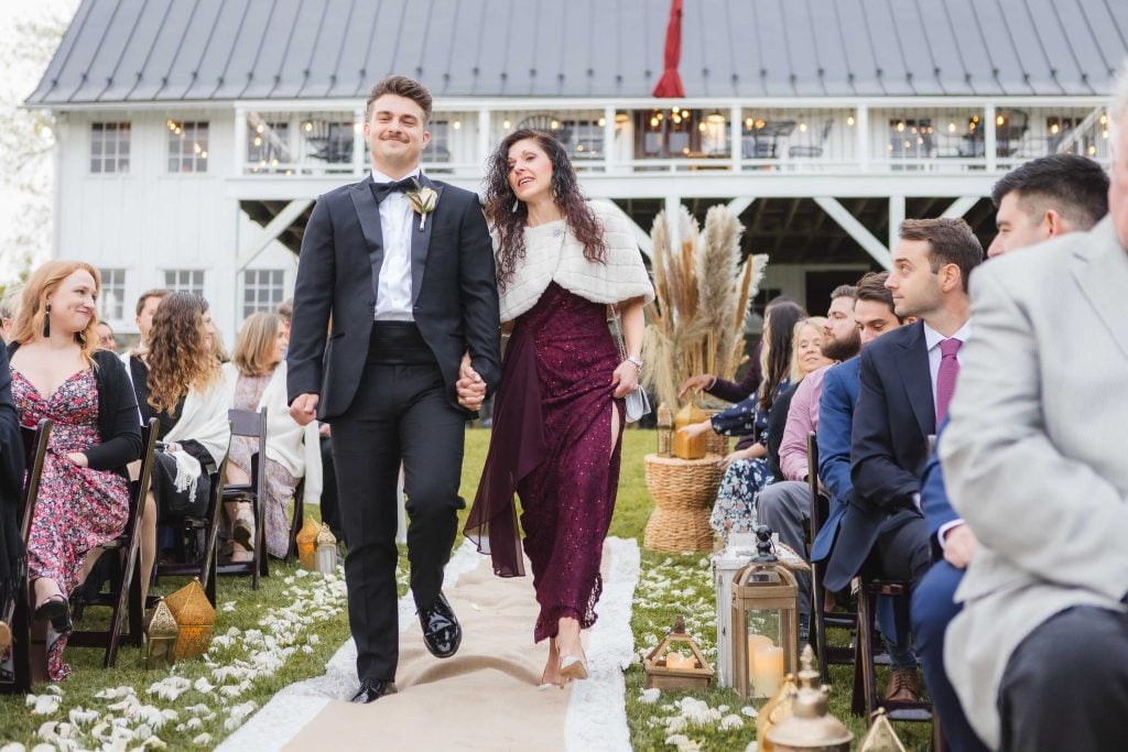 A couple walks down the aisle at an outdoor wedding at The Barns at Hamilton Station, hand in hand, smiling, with guests seated on either side.