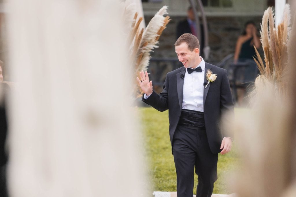 A man in a black tuxedo with a bow tie and boutonniere waves while walking outside at The Barns at Hamilton Station. Pampas grass is in the foreground, and a few blurred figures are in the background, capturing the essence of a vineyard wedding.