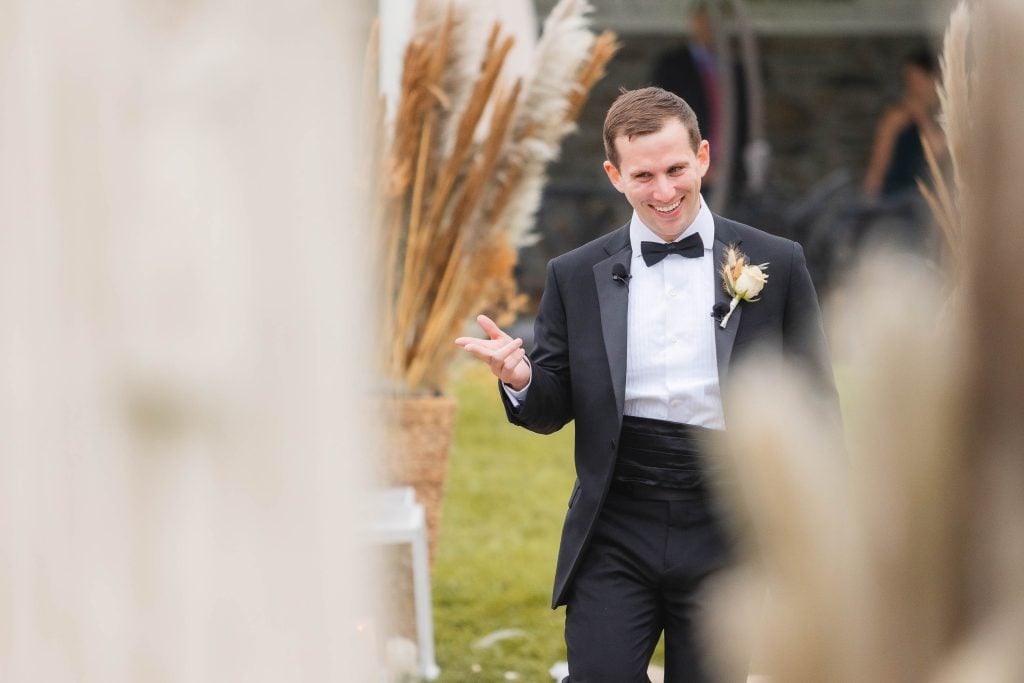 A man in a black tuxedo with a bow tie and boutonniere gestures while standing outdoors at The Barns at Hamilton Station, with decorative plants in the background, capturing the elegance of the wedding ceremony.