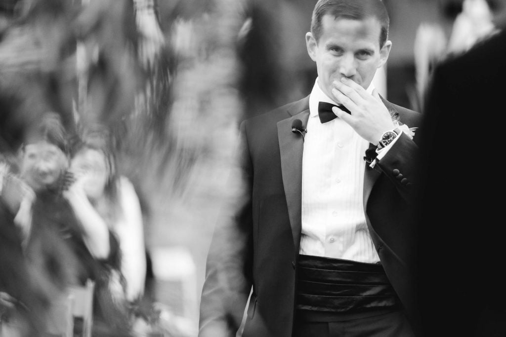 A man in a tuxedo stands outdoors at a vineyard, covering his mouth with his hand. People sitting in the background are blurred, capturing the intimate moment of the ceremony.